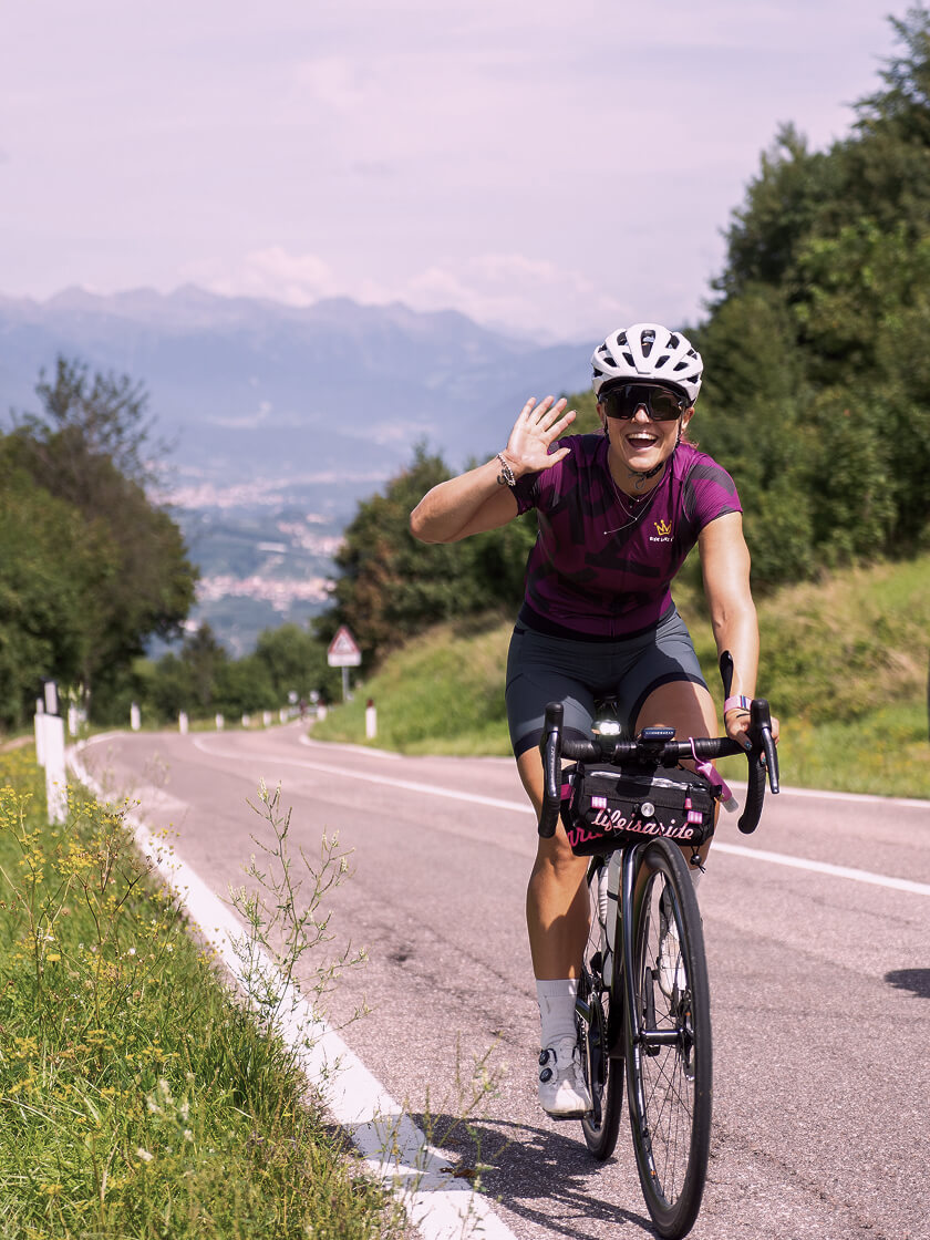 Frau fährt auf Rennrad bergauf, winkt und lacht
